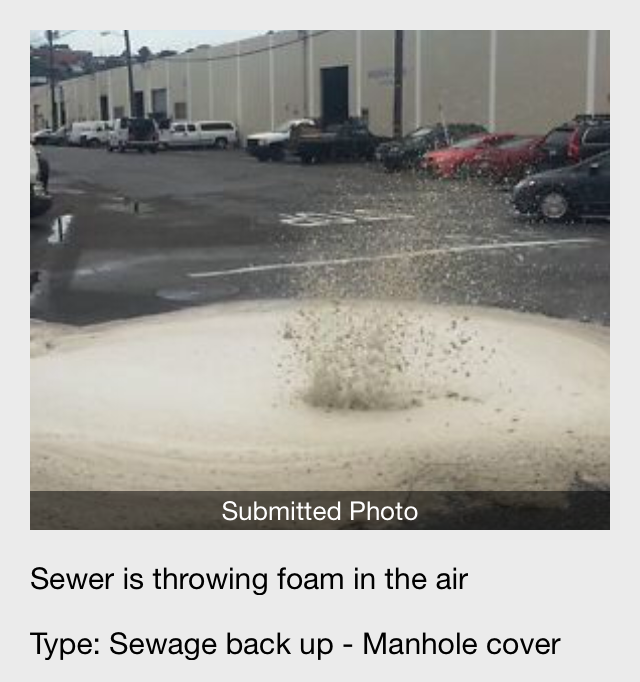 A geyser of white foam blasting out of a sewer in an urban area.